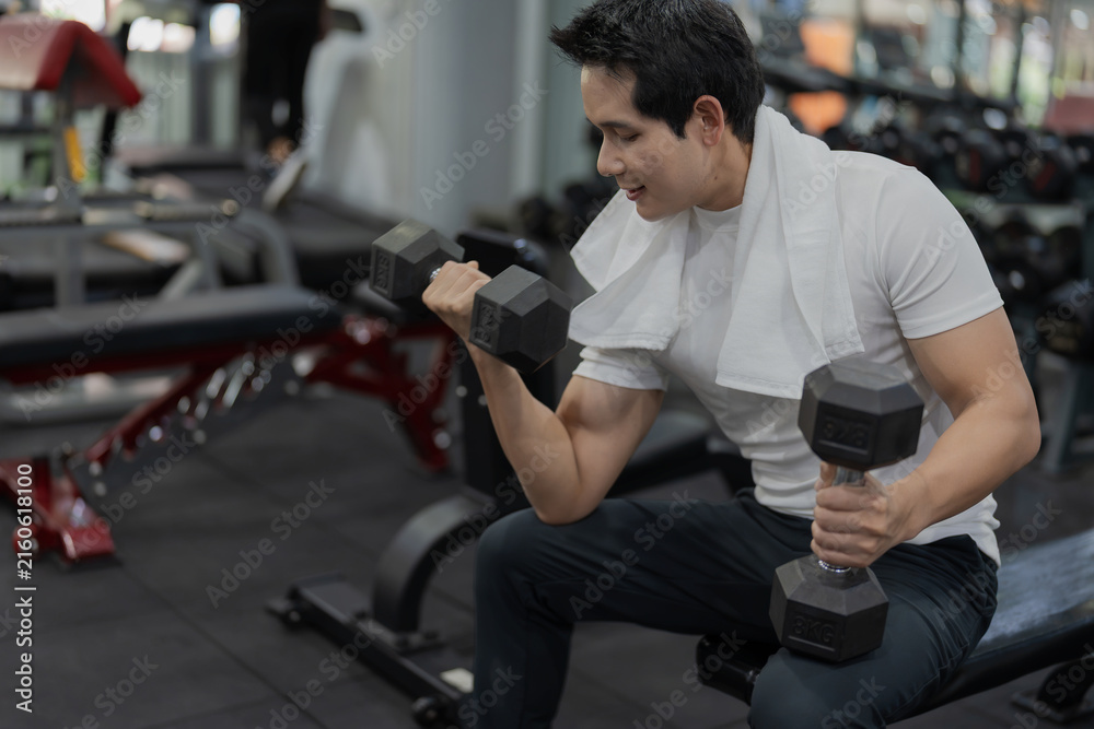 Fototapeta premium Asian man sitting on workout bench doing bicep curls with dumbbells in gym