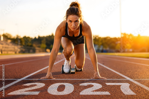 Determined female athlete in starting position on running track with 2027 marking, ready to race at sunset