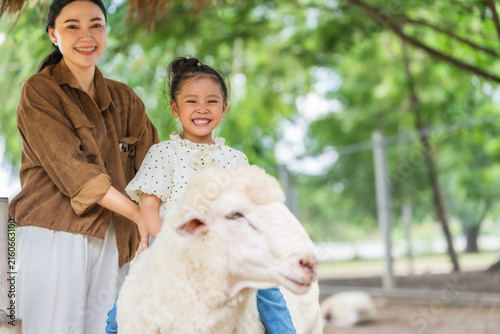 happy preschool child girl riding a sheep with mother beside in outdoor farm