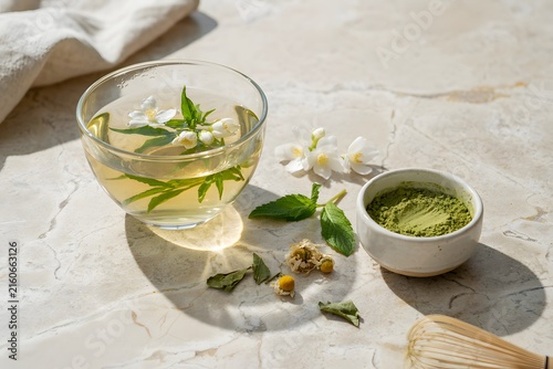 A refreshing glass of jasmine green tea with fresh mint and chamomile flowers, served with matcha powder and a whisk on a marble surface.