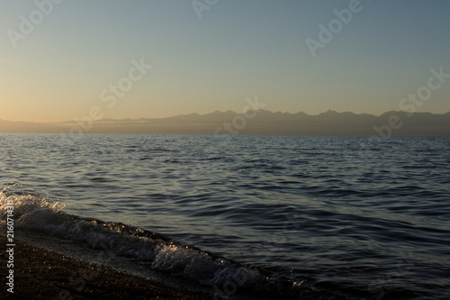 Photography Serene coastline at dawn with gentle waves lapping the shore and distant mountain range silhouetted against a clear sky under soft golden sunlight