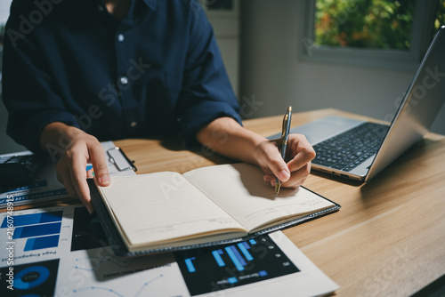 Person hands writing in a notebook, working on business data with charts and a laptop on a wooden desk