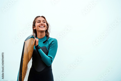 A joyful young woman wearing a teal wetsuit stands outdoors holding a surfboard, smiling brightly during her summer surfing holiday at the beach with a clean sky background.