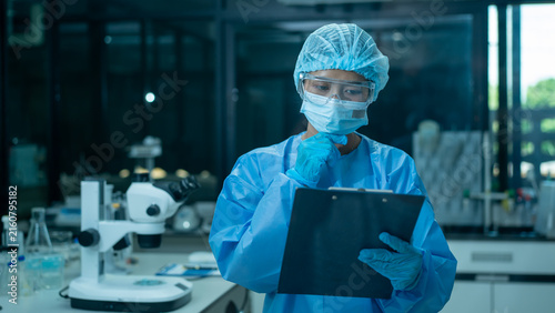 Scientist in protective gear taking notes on a clipboard in a modern laboratory with microscope and glassware in the background
