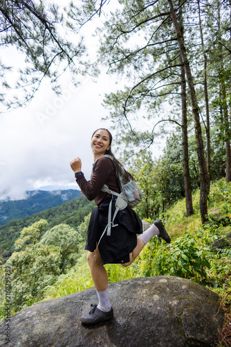 And keywordsSmiling Asian female hiker wearing backpack and standing on forest boulder with mountain landscape, pine tree, and lush vegetation during outdoor adventure