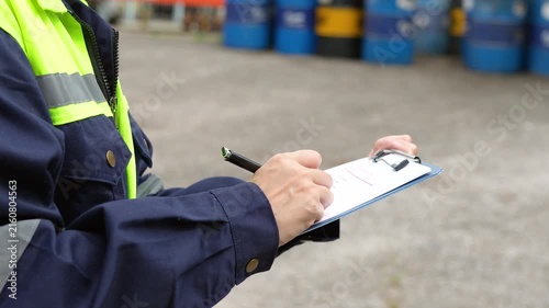 Checking on chemical hazardous safety checklist form, close-up at worker hand, with factory warehouse and metal barrel storage as background. Industrial safe work practice scene, selective focus.