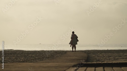 Silhouette of young woman with beach accessories spinning on seashore near wooden walkways of sandy beach. Evening freedom and enjoyment of nature and rest in backlight of Mediterranean coast.