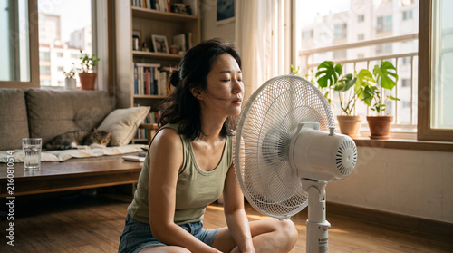 Young woman cooling with electric fan. Female relaxing at home during hot summer weather.