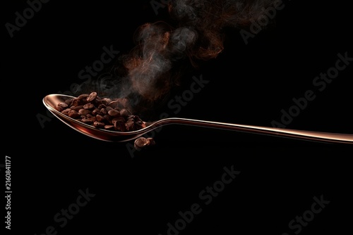A metallic spoon holds roasted beans, releasing fragrant steam against darkness