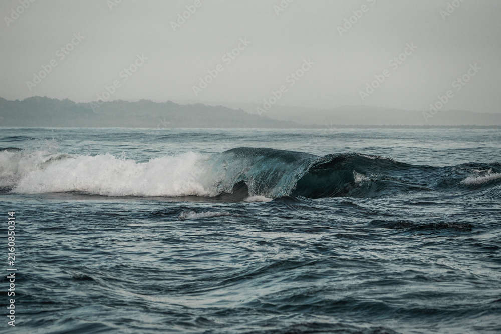 Naklejka premium Misty seascape of rolling teal waves breaking along a distant hazy coastline under an overcast gray sky