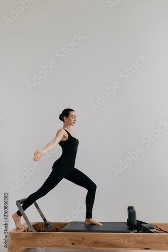 Woman in a black bodysuit performing a barefoot lunge exercise standing on a wooden pilates reformer machine, arms extended forward for balance and core control inside a bright minimalist studio room