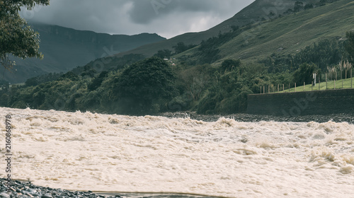 Dramatic mountain river overflowing with powerful muddy rapids during severe flood beneath cloudy green hillside landscape