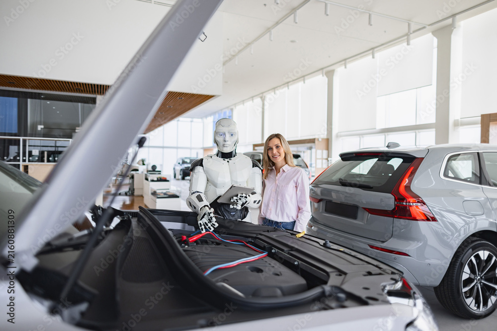 Fototapeta premium A humanoid robot with a tablet inspects a car engine while a woman stands nearby in a modern dealership showroom