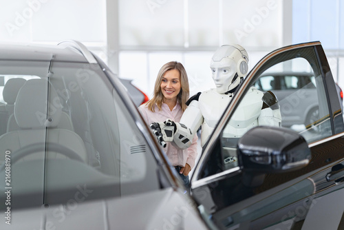 A humanoid robot stands next to a smiling woman, gesturing towards a car in a modern dealership showroom