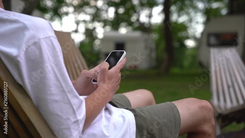 Person lounges in chair, scrolling phone outdoors. Relaxed posture, focused on device. Background shows campsite with trailers. Ideal for vacation, travel, or digital lifestyle themes