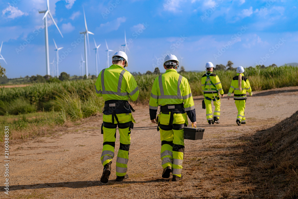 Fototapeta premium A maintenance team walks away down a long dirt track that runs between rows of wind turbines, the leading pair close together and two more figures following far behind