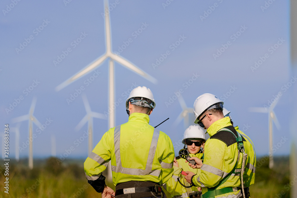 Fototapeta premium A small wind farm crew checks a handheld wind meter together in the late light, one man's arm crossing the frame to hold the probe while a colleague watches from behind