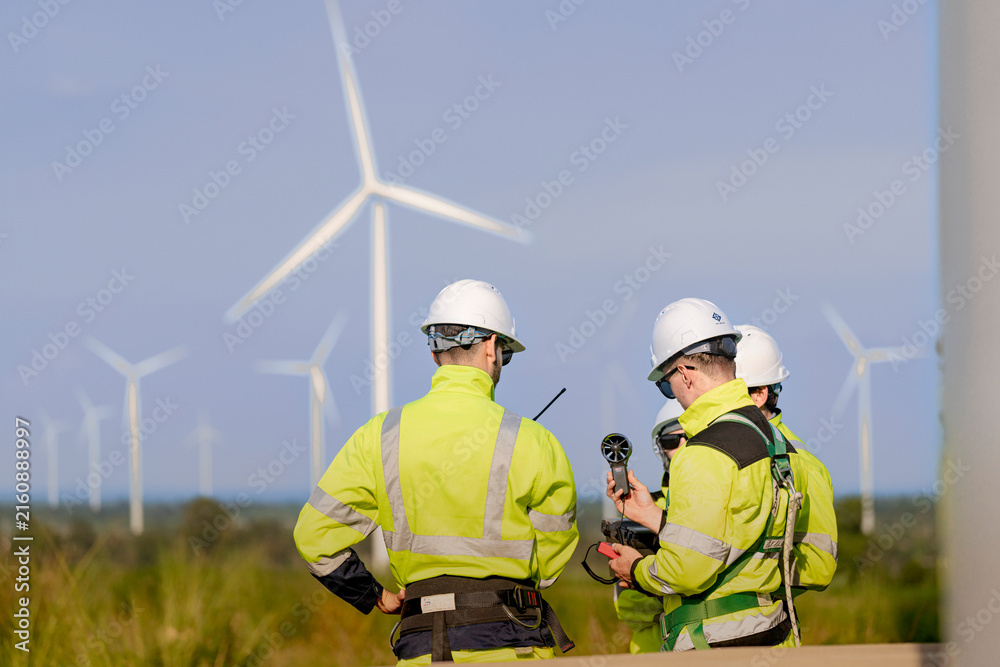 Fototapeta premium A wind technician holds a vane anemometer at chest height while his teammate leans in from the right, both framed against a bright sky with turbines soft in the background