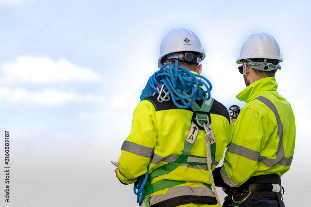 Fototapeta premium Seen from behind, two rope access technicians stand together on the wind farm with a large blue rope coil between them, their white hard hats bright against the pale sky