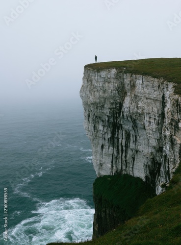 Lone figure stands on the edge of a sheer cliff overlooking a turbulent sea
