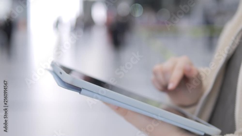 Person holds tablet, taps screen in public space. Background shows blurred people walking. Hand gestures suggest digital interaction. Modern tech used for communication or work