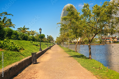Fotografía Pedestrian stone footpath receding into distance in green urban park near waterfront lake with modern skyscrapers, trees, palm foliage, lawn, lanterns under blue summer sky
