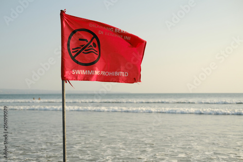 Red Warning Flag Fluttering Against the Ocean with a No Swimming Sign, Beach Safety and Hazard Concept