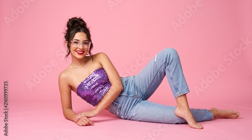 Radiant young woman with a high bun hairstyle wearing a sparkling purple sequin tube top and blue denim jeans poses gracefully for a vibrant portrait.