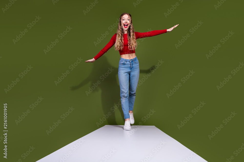 Naklejka premium Young woman in red top and jeans balances on a white platform with green background
