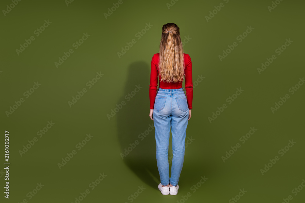 Fototapeta premium Young woman standing with red top and jeans facing away against green background