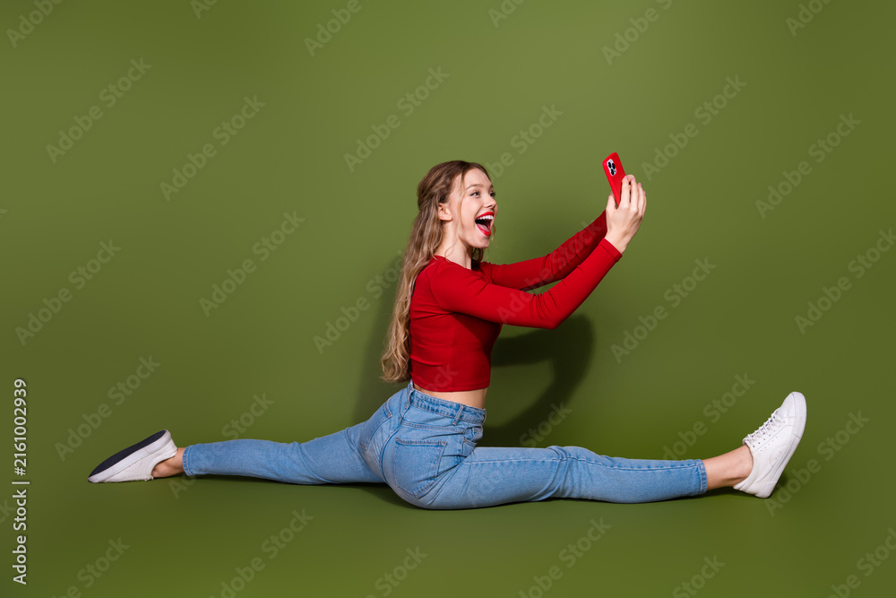 Naklejka premium Young woman performs a full split while taking a selfie in a bright red top and jeans against a green backdrop