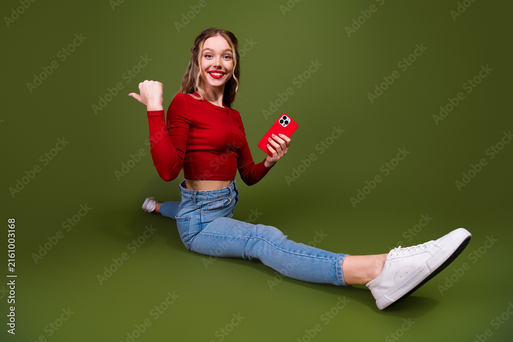 Naklejka premium Young woman smiling sits on green studio background wearing red top jeans holding red phone for lifestyle advertising