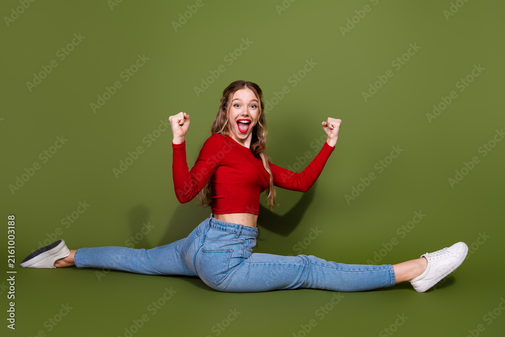 Naklejka premium Young woman performs a full split in red top jeans against green backdrop smiling and energetic