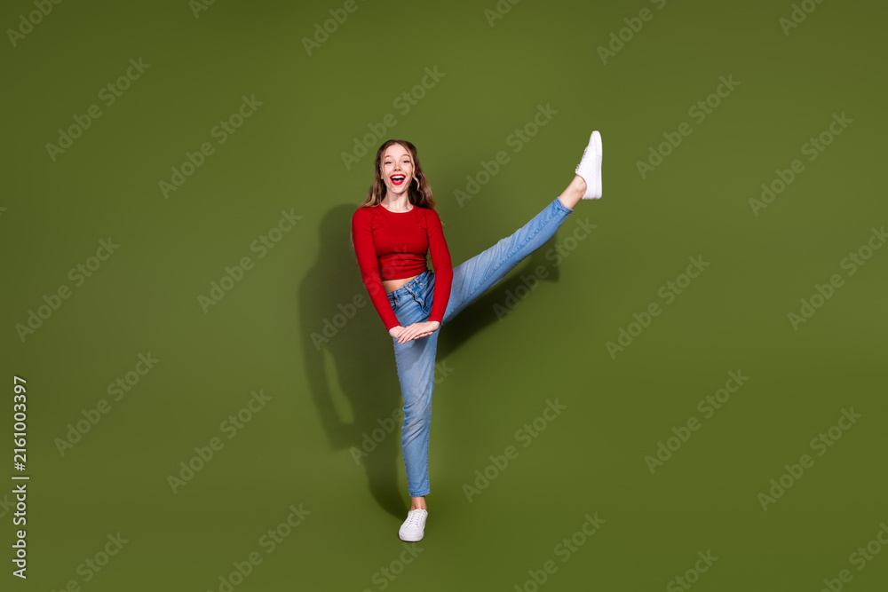 Naklejka premium Young woman in red top and jeans kicks her leg up on green studio backdrop smiling