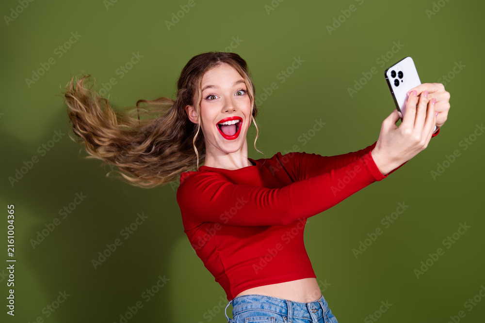 Naklejka premium Young woman taking a selfie in a red top smiling with phone against green background