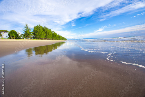 Beautiful tropical beach landscape during a sunny daytime, featuring clear blue skies, calm ocean water, sandy shoreline, and natural coastal scenery. Bright sunlight creates a peaceful and relaxing a