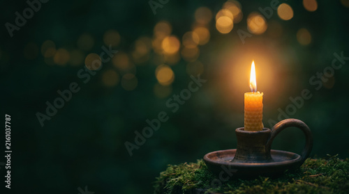Burning beeswax candle in vintage holder on green moss with warm bokeh lights and dark copy space