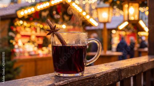 A glass of mulled wine with a cinnamon stick and star anise sits on a wooden railing. Festive lights and market stalls are blurred in the background.