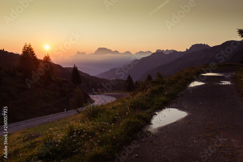 Dolomites mountain peaks and sunrise over a winding road with puddles on a dirt path in the Passo Valles area, Dolomites, Italy
