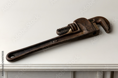 Close-up of an old, rusty pipe wrench resting on a light surface. Shows texture and detail of the vintage tool, a classic piece of hardware.