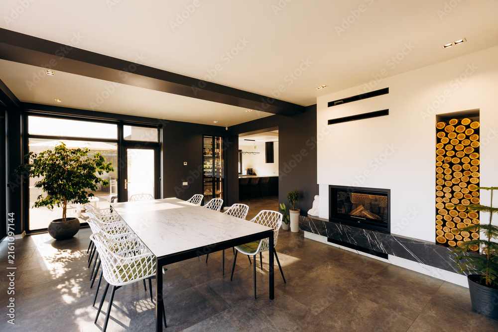 Fototapeta premium Modern dining room with long marble table, black metal chairs, built-in fireplace, stacked firewood, glass doors, indoor plants, open kitchen view and bright neutral decor