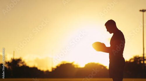 A football player in silhouette looking down at a rugby ball during a sunset, capturing a moment of intense focus, reflection, and quiet preparation for the game.