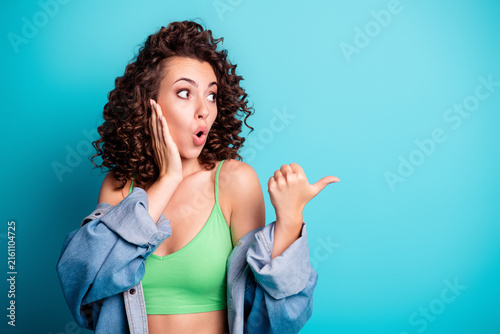 Fotografie Surprised young woman with curly hair in green top and denim jacket pointing to