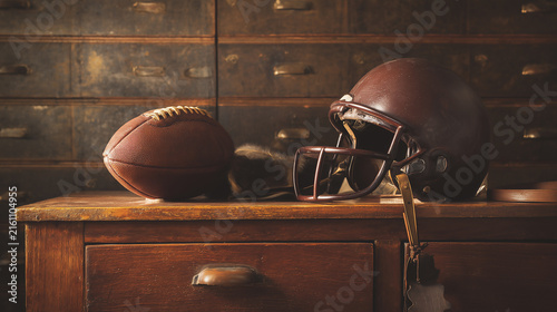 A weathered American football helmet and a classic leather football sit on a rustic wooden table in front of old storage drawers, evoking the nostalgic spirit of vintage equipment and history