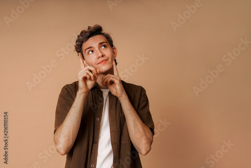 Thoughtful young man looking up with thinking gesture on beige background
