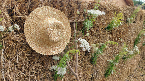 Photography Traditional straw hat hanging on wooden rake against hay bale wall decorated wit