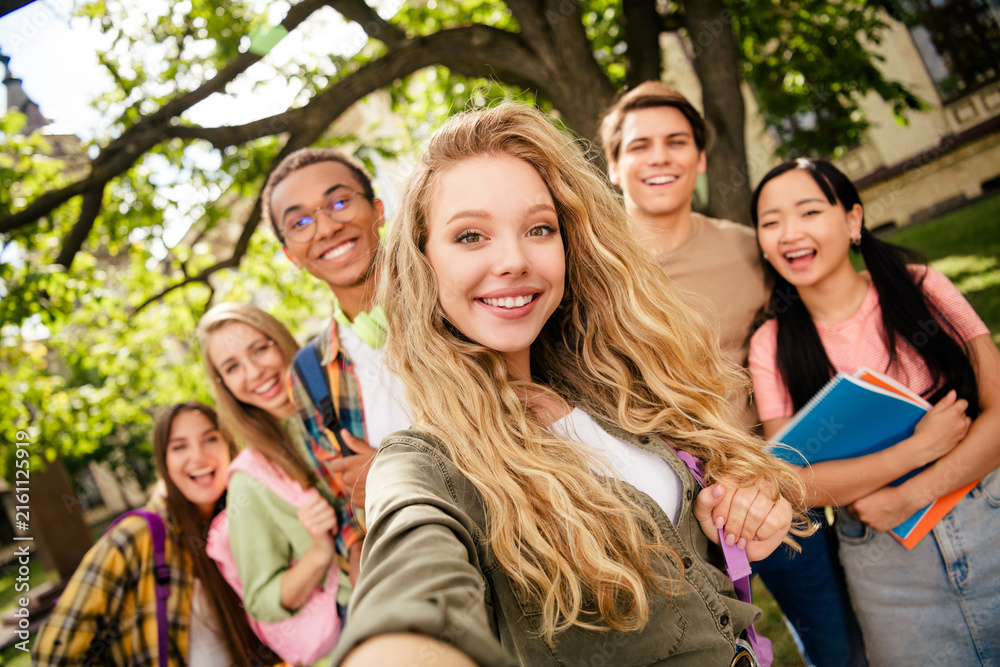 Fototapeta premium Group of smiling college students taking a campus selfie together, diverse young friends enjoying outdoor university life and studying with notebooks