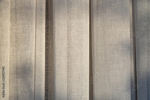Closeup of textured light vertical folds of translucent curtain fabric in soft sunlight with shadows.