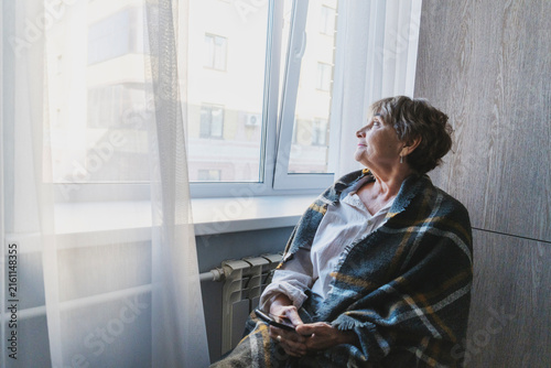 Lonely senior Caucasian woman sitting by a radiator near the window, wrapped in a blanket with a smartphone, conceptualizing pensioner isolation.