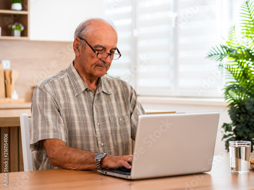 Senior man using laptop at home, sitting at the table in the living room and concentrating on work with serious and focused expression.